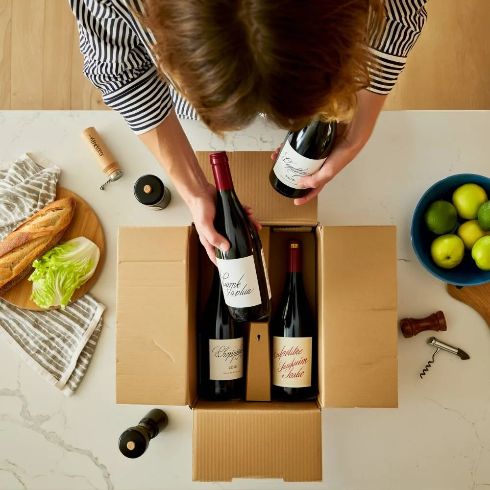 Person packing wine bottles into a cardboard box on a kitchen counter.