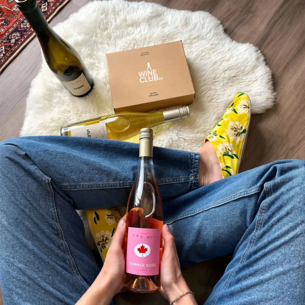 Person holding a bottle of wine with a Wine Club box and another bottle on a rug.