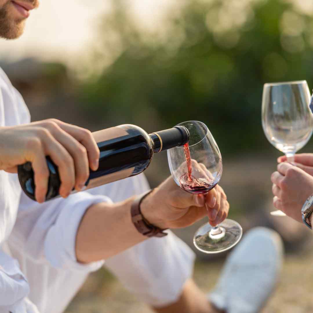 A bearded man well dressed serving red wine in glass in a vineyard